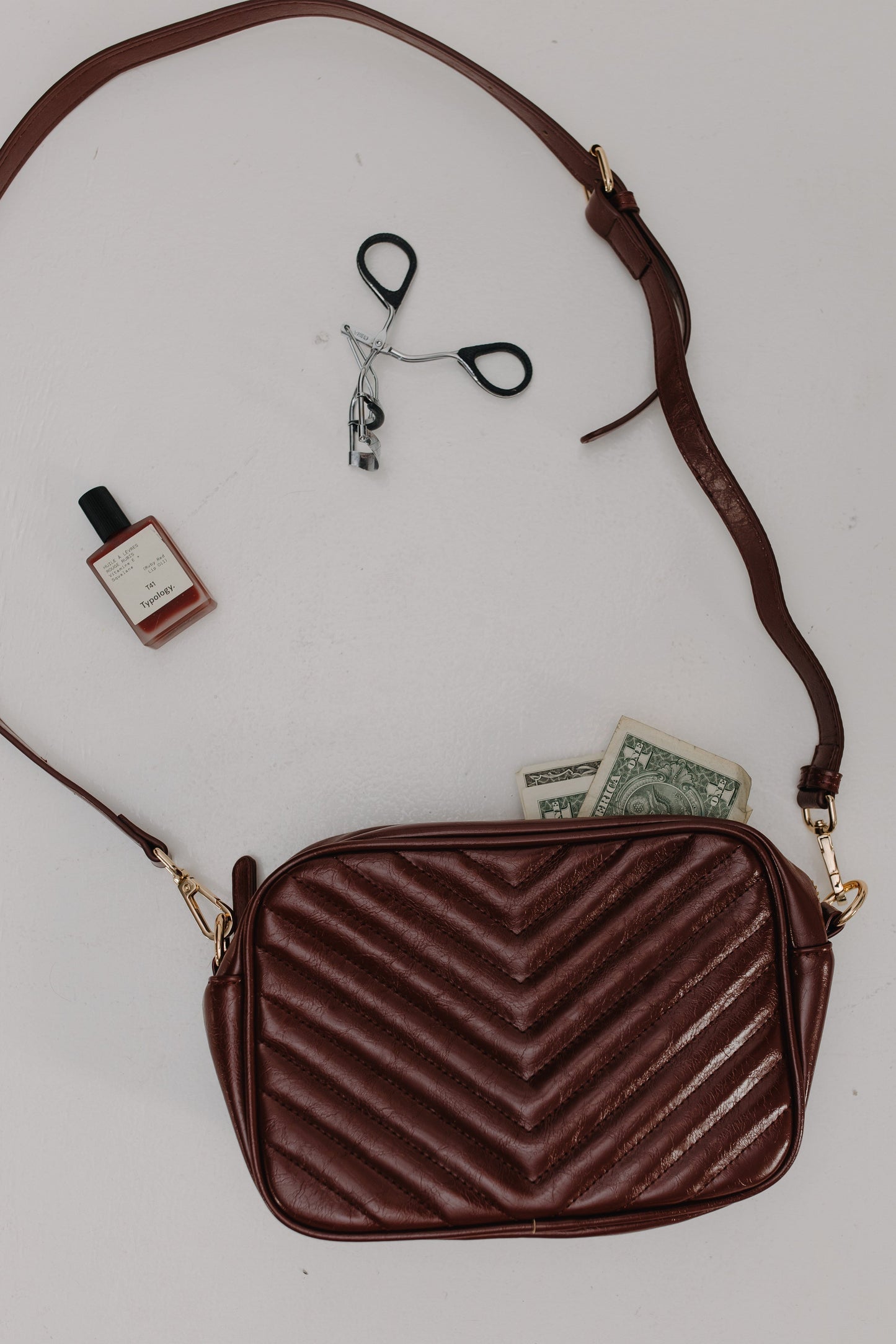 Burgundy chevron-patterned handbag with a strap, small bottle, and money on a light gray background