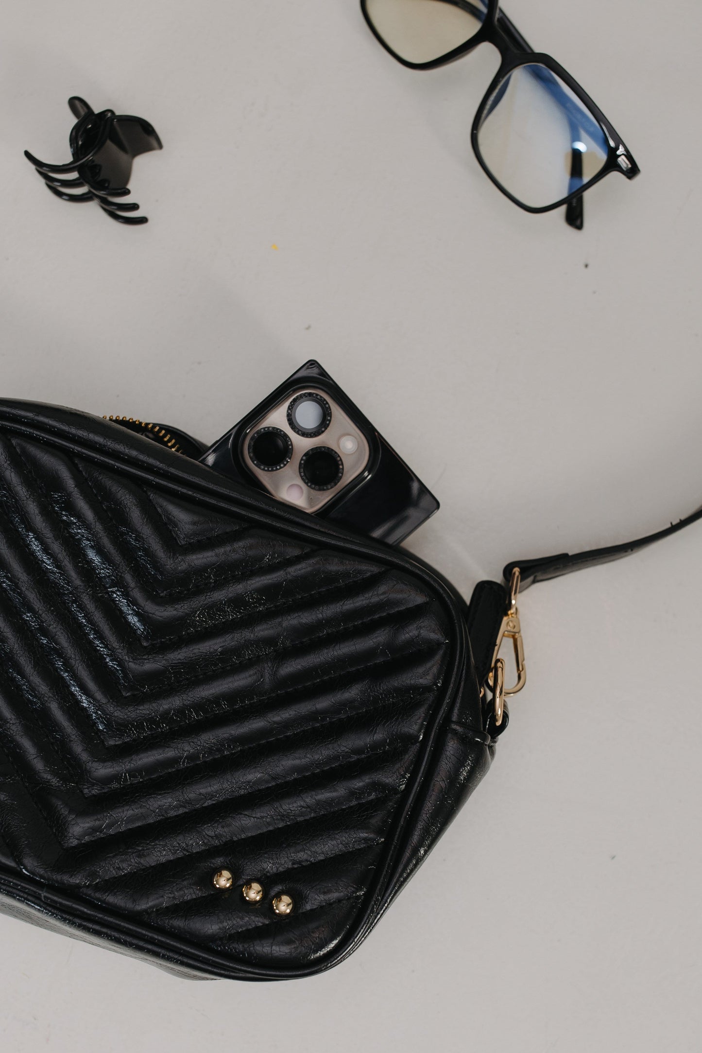 Black quilted handbag with a phone, glasses, and small black spider toy on a light gray background