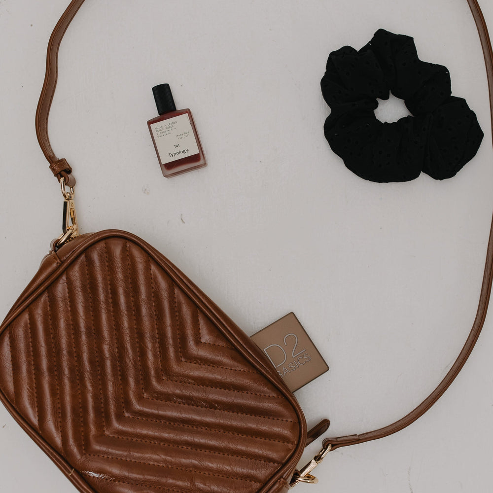 Brown textured crossbody bag with a black scrunchie and small bottle on a white background