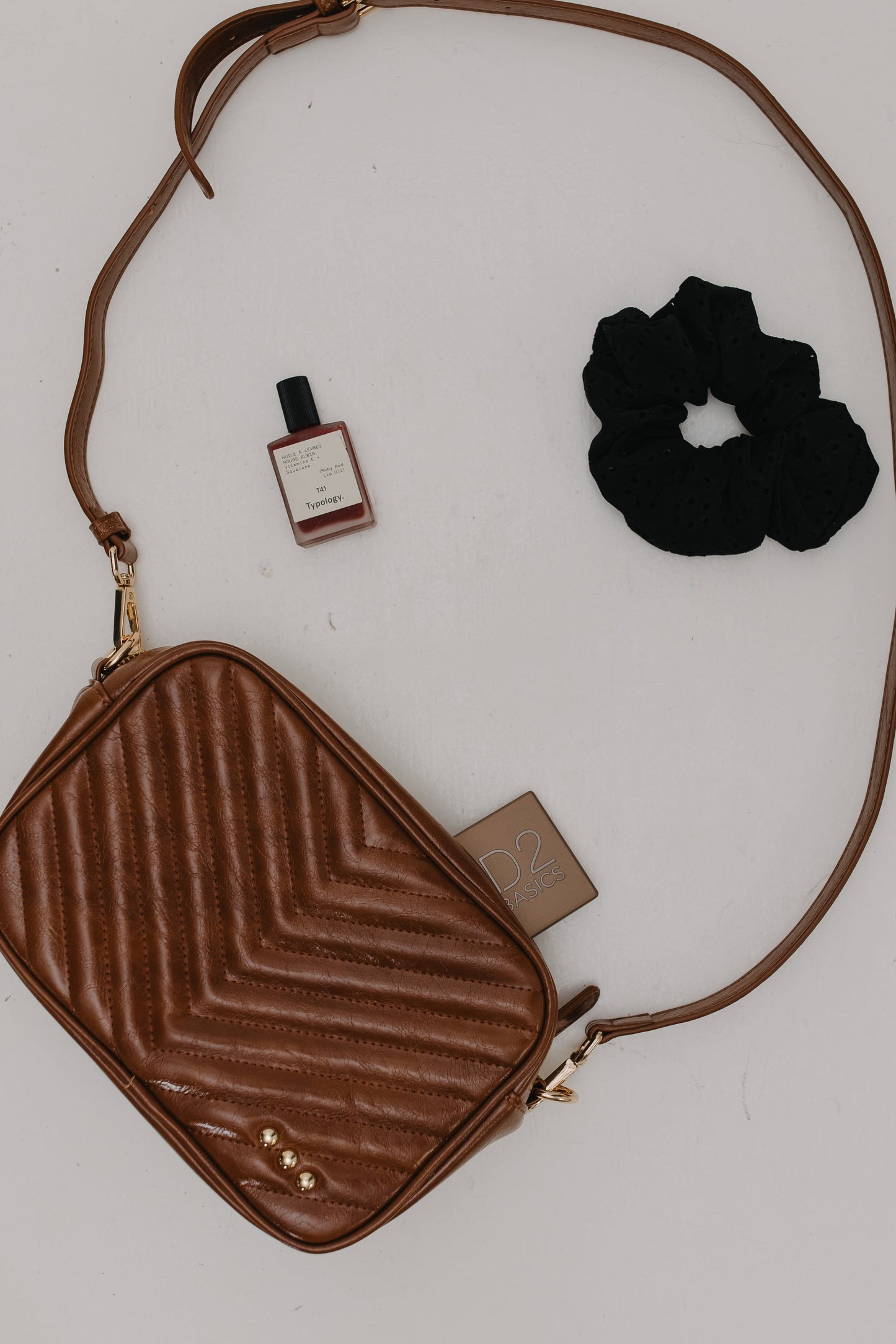 Brown textured crossbody bag with a black scrunchie and small bottle on a white background
