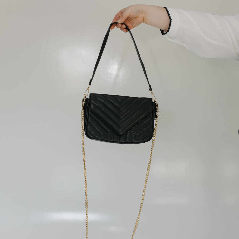 Black quilted handbag with a gold chain strap held by a person against a light gray background