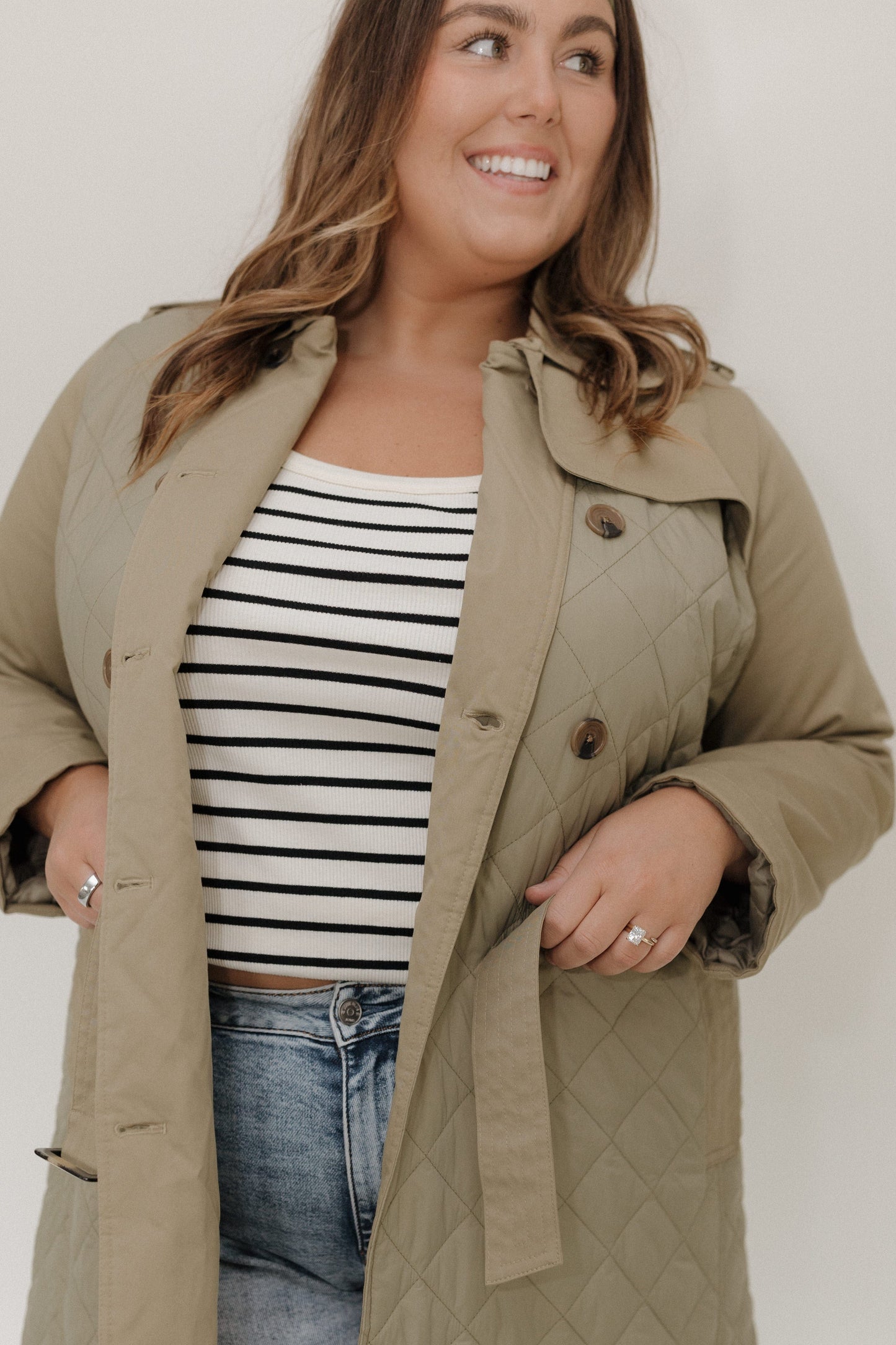 Woman wearing a beige trench coat over a striped shirt and jeans against a plain background