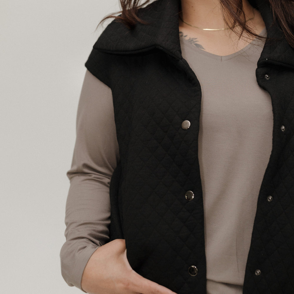 Woman wearing a black quilted vest over a beige top against a plain background
