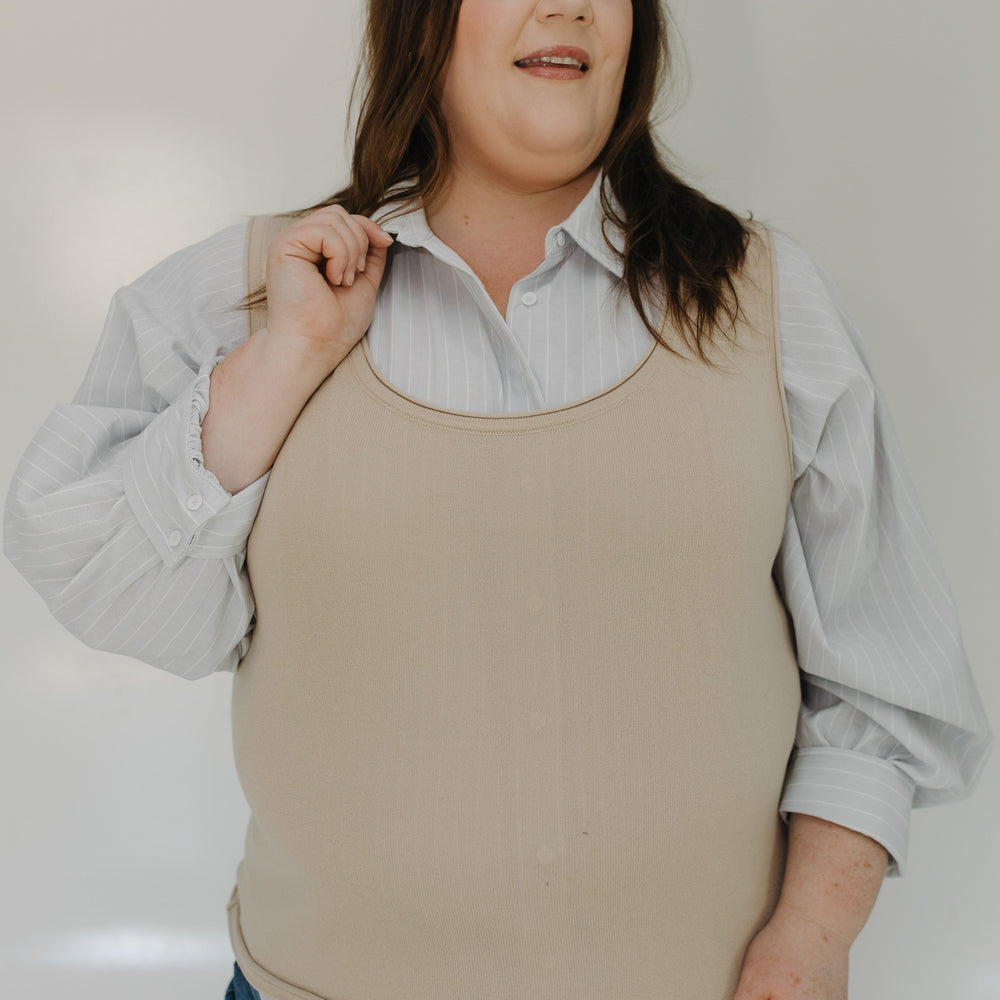 Woman wearing a beige sleeveless top over a light blue shirt with rolled-up sleeves, paired with blue jeans.
