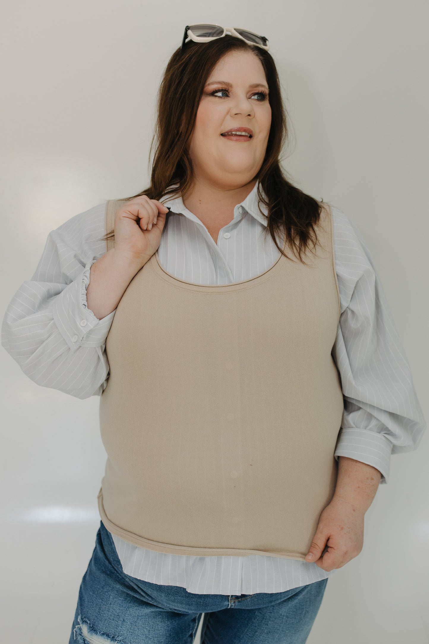 Woman wearing a beige sleeveless top over a light blue shirt with rolled-up sleeves, paired with blue jeans.
