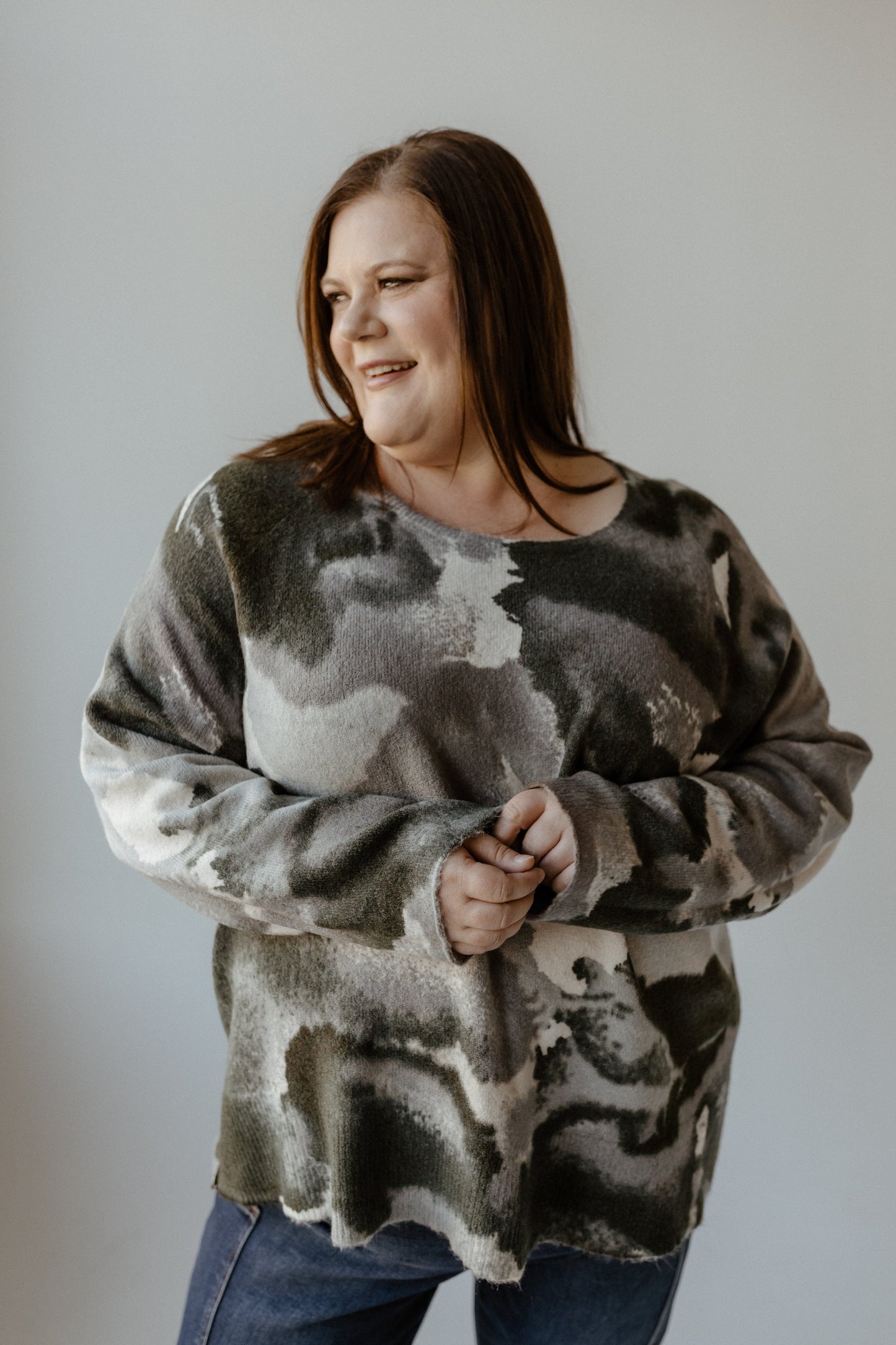 A person wearing a versatile, camouflage-patterned sweater and jeans stands against a plain background, smiling while looking to the side.