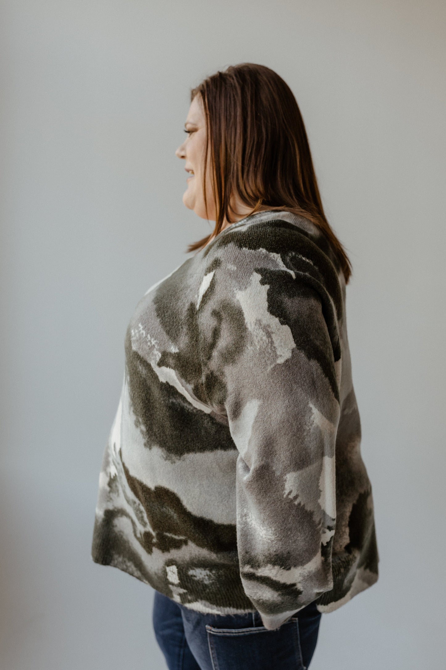 A person with long hair stands in profile, wearing a tunic-length camouflage-patterned sweater and jeans, set against a plain background.