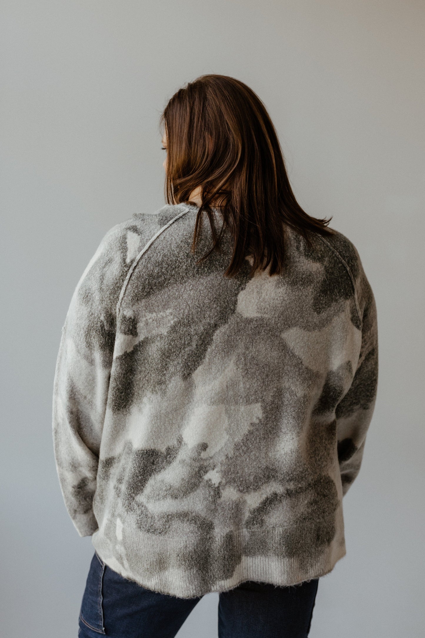 A person wearing a versatile, gray and white camouflage-patterned sweater stands facing away. Their long brown hair flows gracefully against the plain background, perfectly complementing the tunic-length design of the sweater.