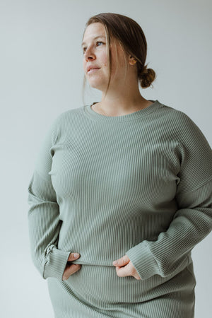 A person wearing a Mikarose ribbed cropped sweater in faded jade with dolman sleeves stands with hands in pockets, looking away from the camera.