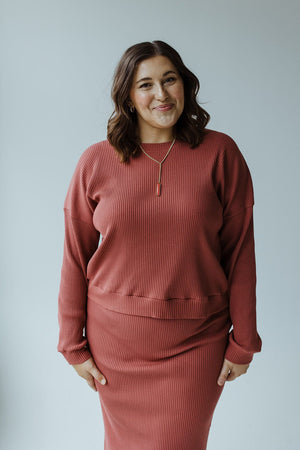 A person wearing a RIBBED CROPPED SWEATER IN SMOKEY ROSEWOOD by Mikarose stands against a plain backdrop, smiling gently.