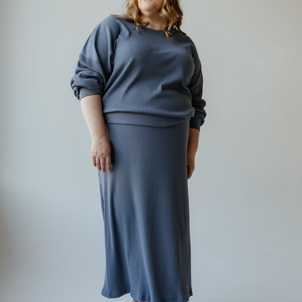 A person wearing the Mikarose Ribbed Knit Midi Length Pencil Skirt in Adrift, featuring long sleeves and an adjustable drawstring waist, stands against a neutral background, looking to the side with a relaxed posture.