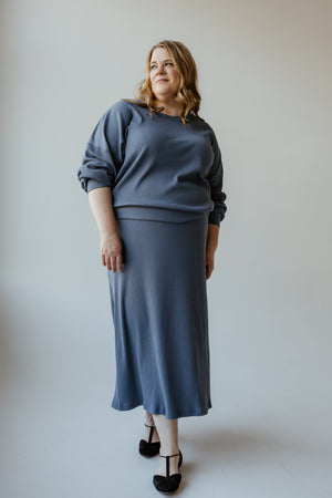 A person wearing the Mikarose Ribbed Knit Midi Length Pencil Skirt in Adrift, featuring long sleeves and an adjustable drawstring waist, stands against a neutral background, looking to the side with a relaxed posture.