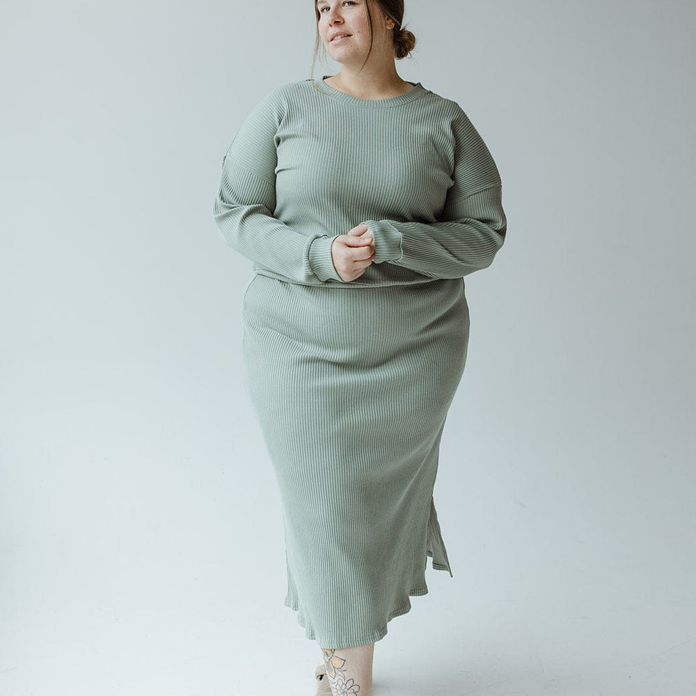 A woman stands in a studio against a plain background, wearing the Ribbed Knit Midi Length Pencil Skirt in Faded Jade by Mikarose and brown sandals. She has light hair styled in a low bun and has a tattoo on her right ankle.