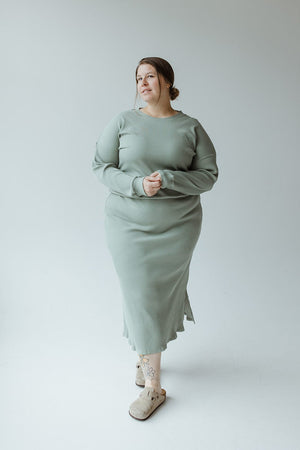 A woman stands in a studio against a plain background, wearing the Ribbed Knit Midi Length Pencil Skirt in Faded Jade by Mikarose and brown sandals. She has light hair styled in a low bun and has a tattoo on her right ankle.