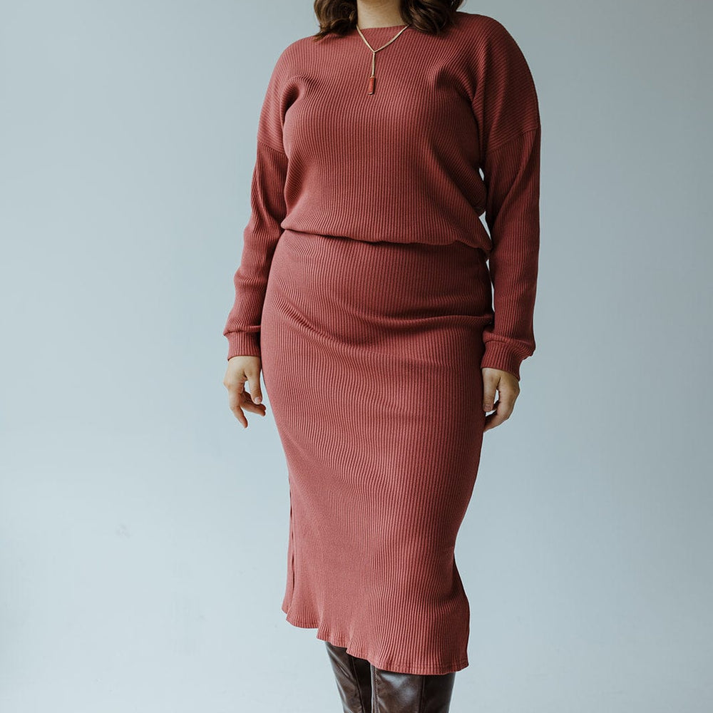 A woman wearing the Mikarose Ribbed Knit Midi Length Pencil Skirt in Smokey Rosewood and a rust-colored sweater, complemented by brown knee-high boots, stands against a plain gray background.