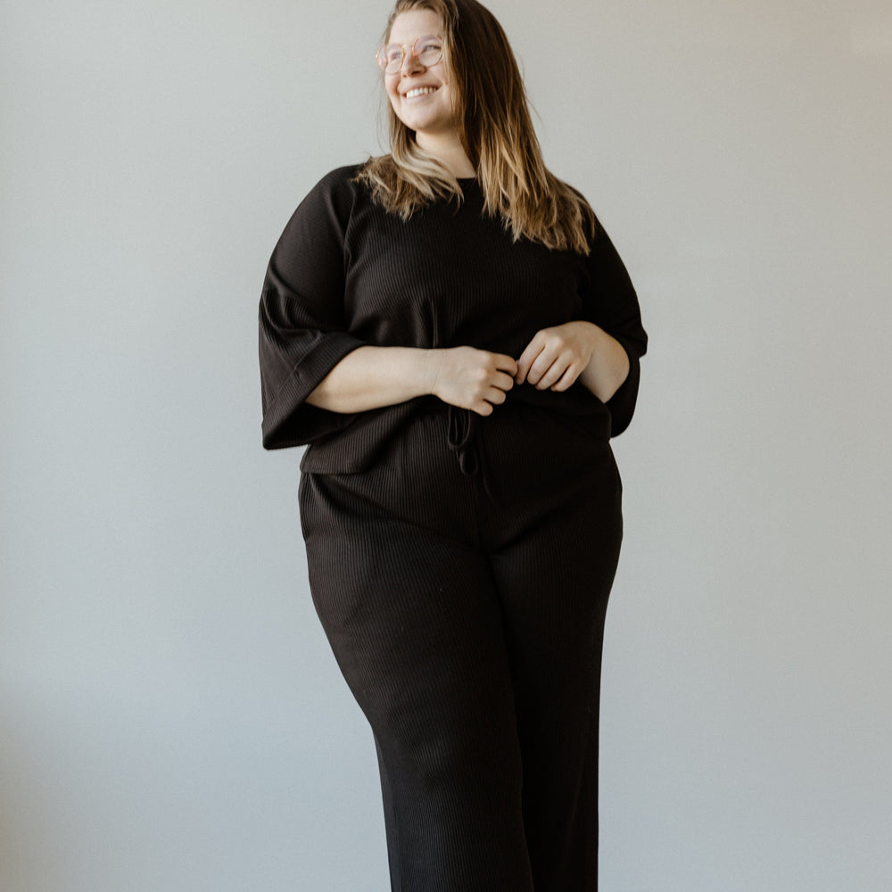 A person in a black outfit, donning stylish high waist pants, is standing in a neutral-toned room, smiling and looking to the side.