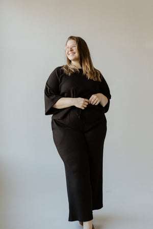 A person in a black outfit, donning stylish high waist pants, is standing in a neutral-toned room, smiling and looking to the side.