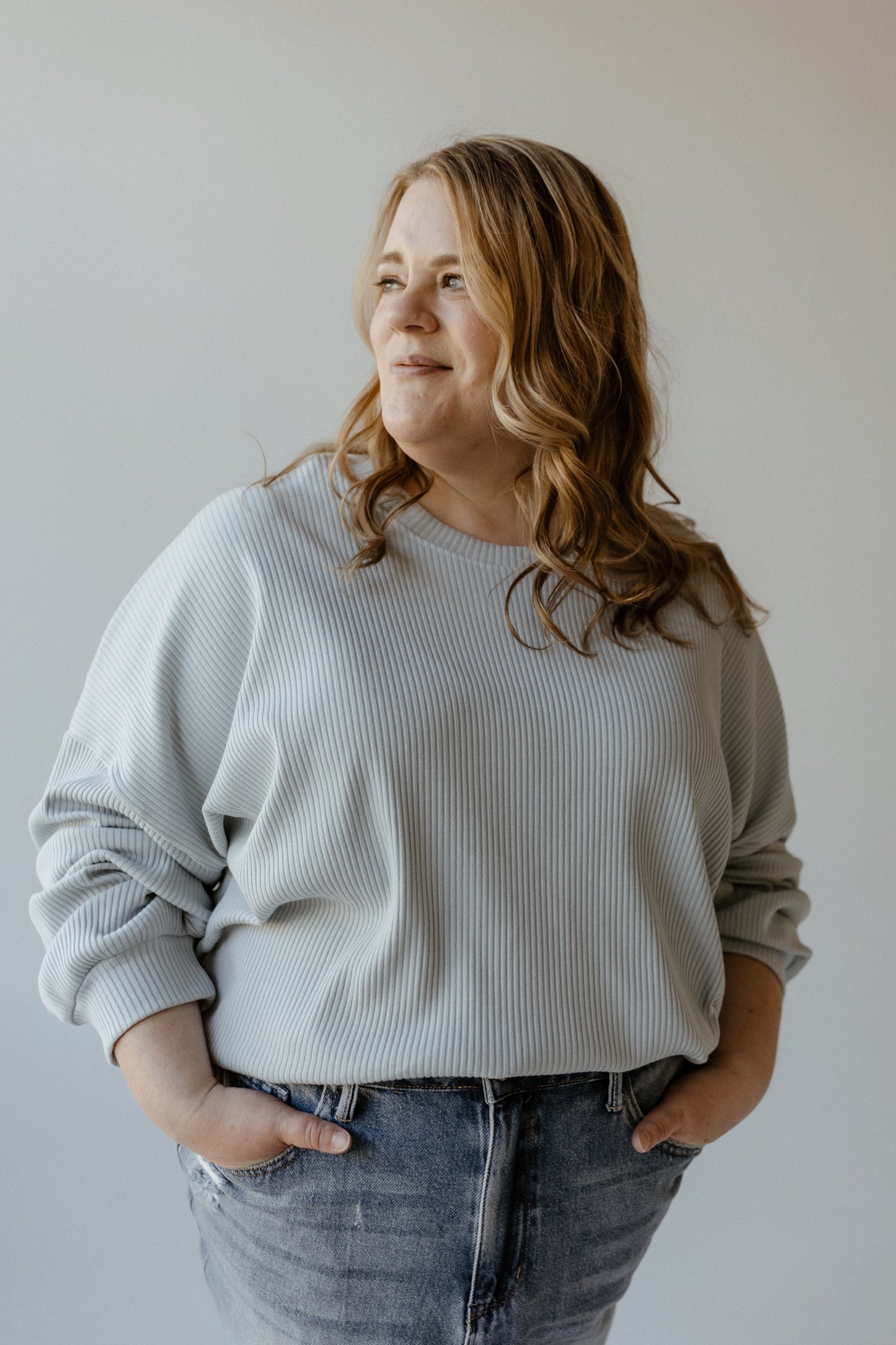 Individual with long hair wearing the Mikarose Ribbed Cropped Sweater in Baby Blue paired with jeans, standing with hands in pockets, gazing to the side against a plain background.