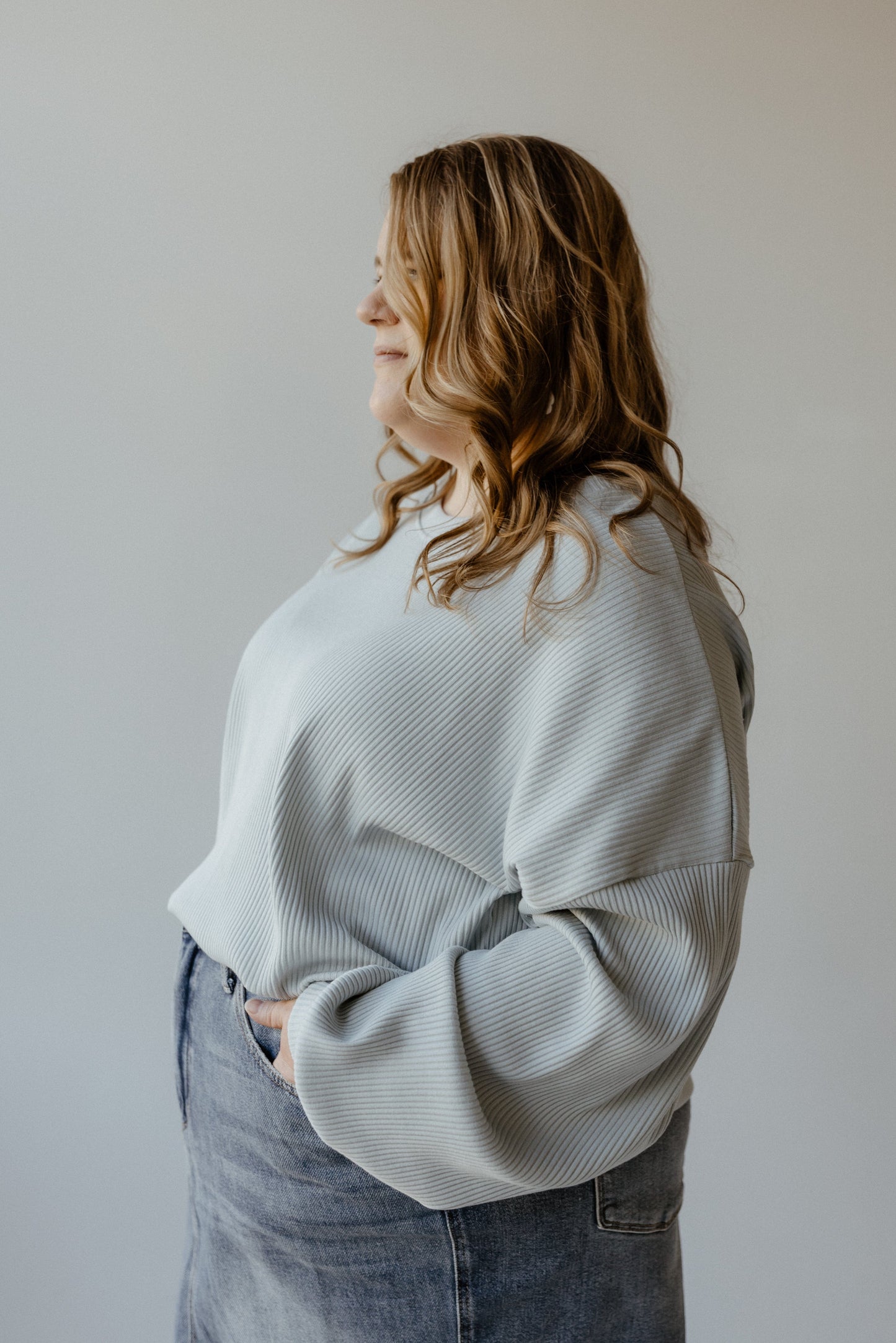 A person with long wavy hair stands sideways, smiling, wearing the Mikarose Ribbed Cropped Sweater in Baby Blue and a denim skirt against a neutral background.