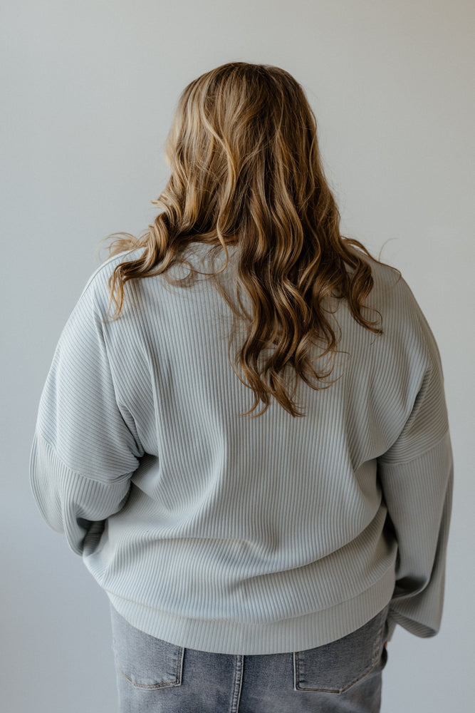 A person with long wavy hair, wearing a ribbed baby blue cropped sweater from Mikarose along with jeans, seen from behind against a plain background.