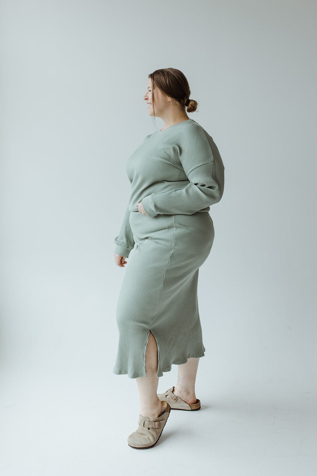 Individual wearing a ribbed cropped sweater in faded jade by Mikarose, paired with beige sandals, posed sideways against a plain background.