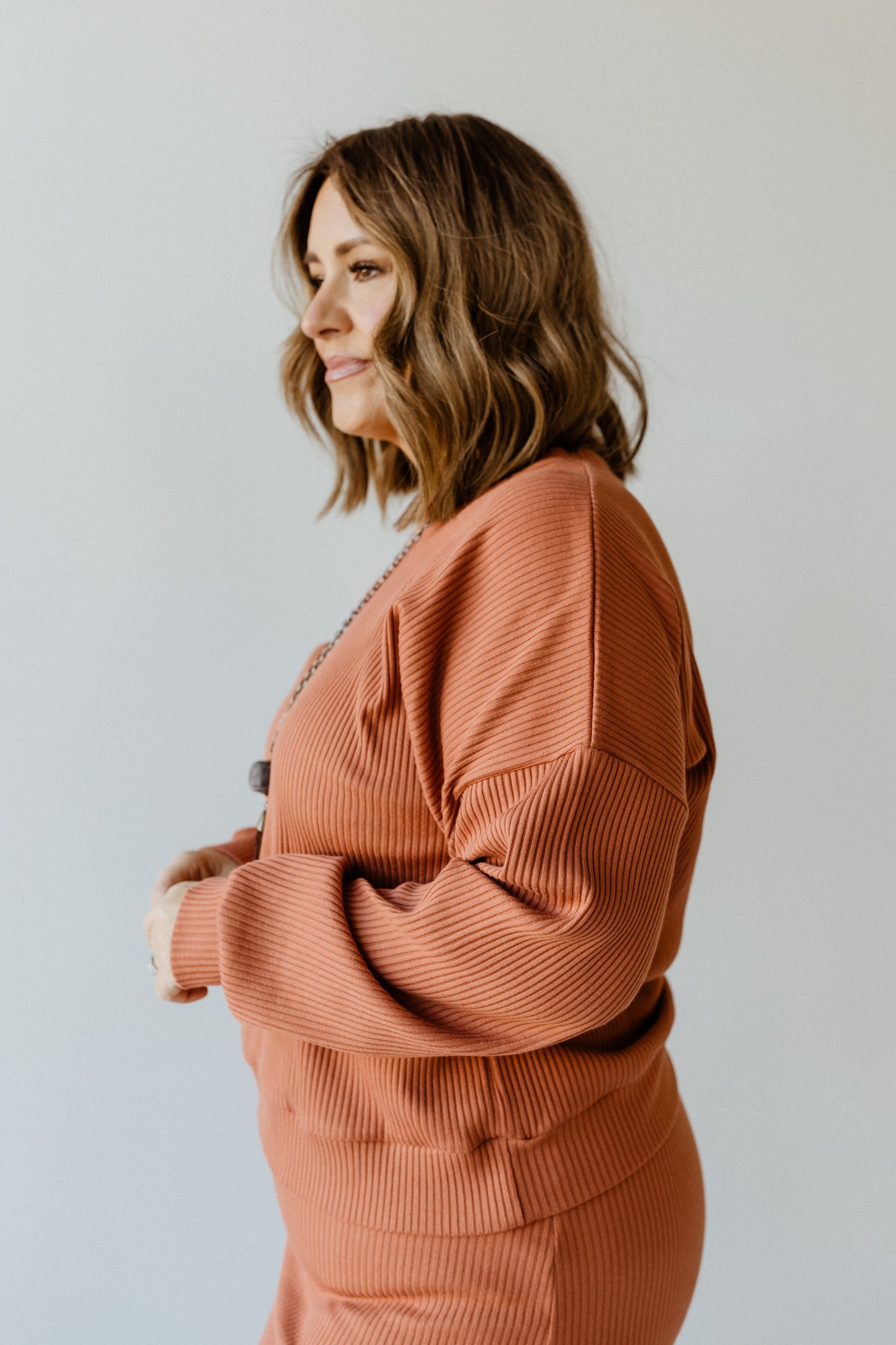 A woman with shoulder-length hair stands in profile, wearing a ribbed cropped sweater and skirt in rusty orange against a plain background.