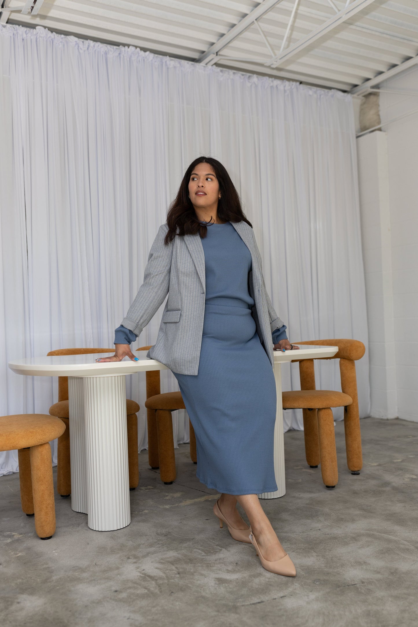 Woman in a blue dress and gray blazer standing in a room with white curtains and wooden furniture.