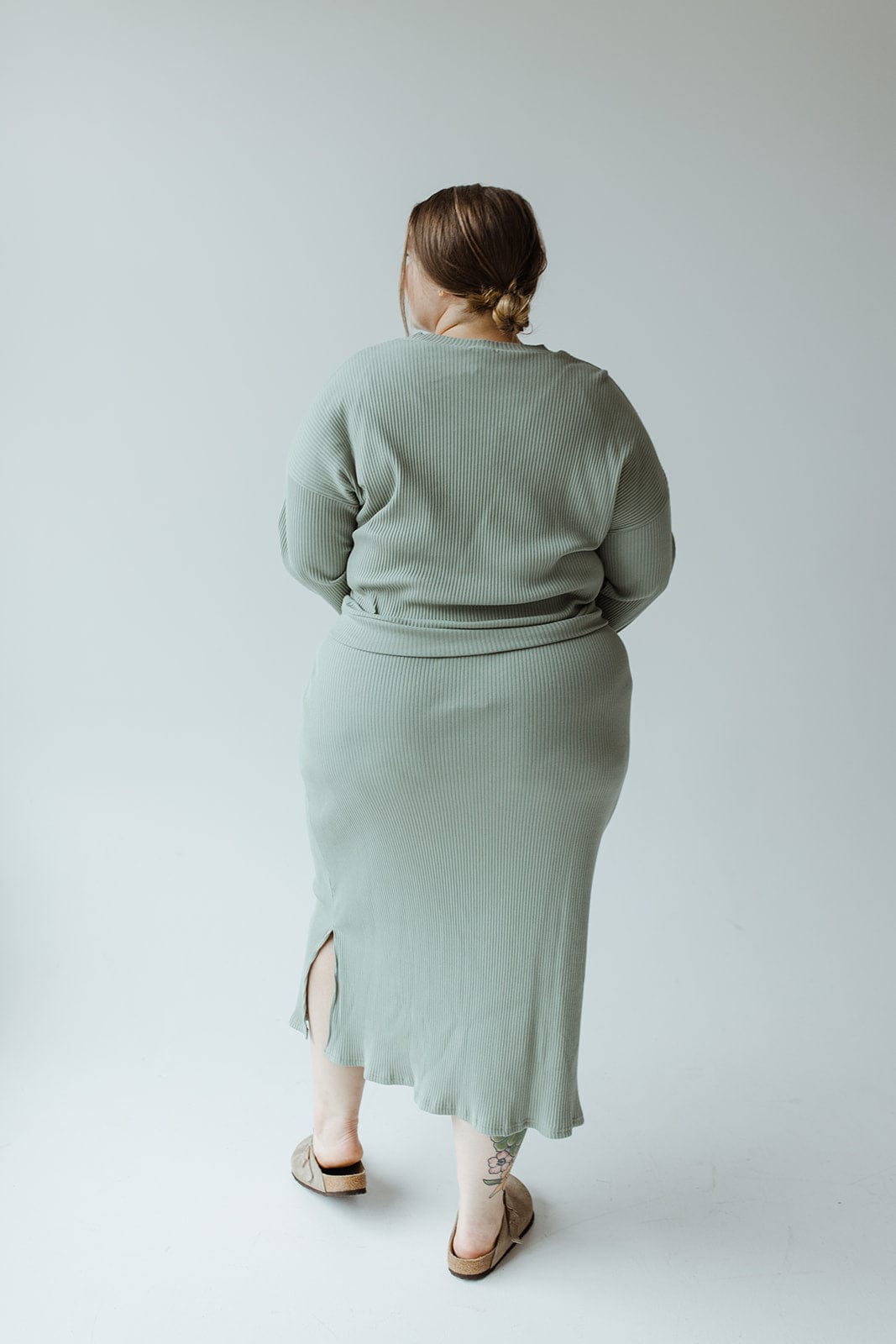 A person stands against a plain background, wearing a light green ribbed sweater and the 