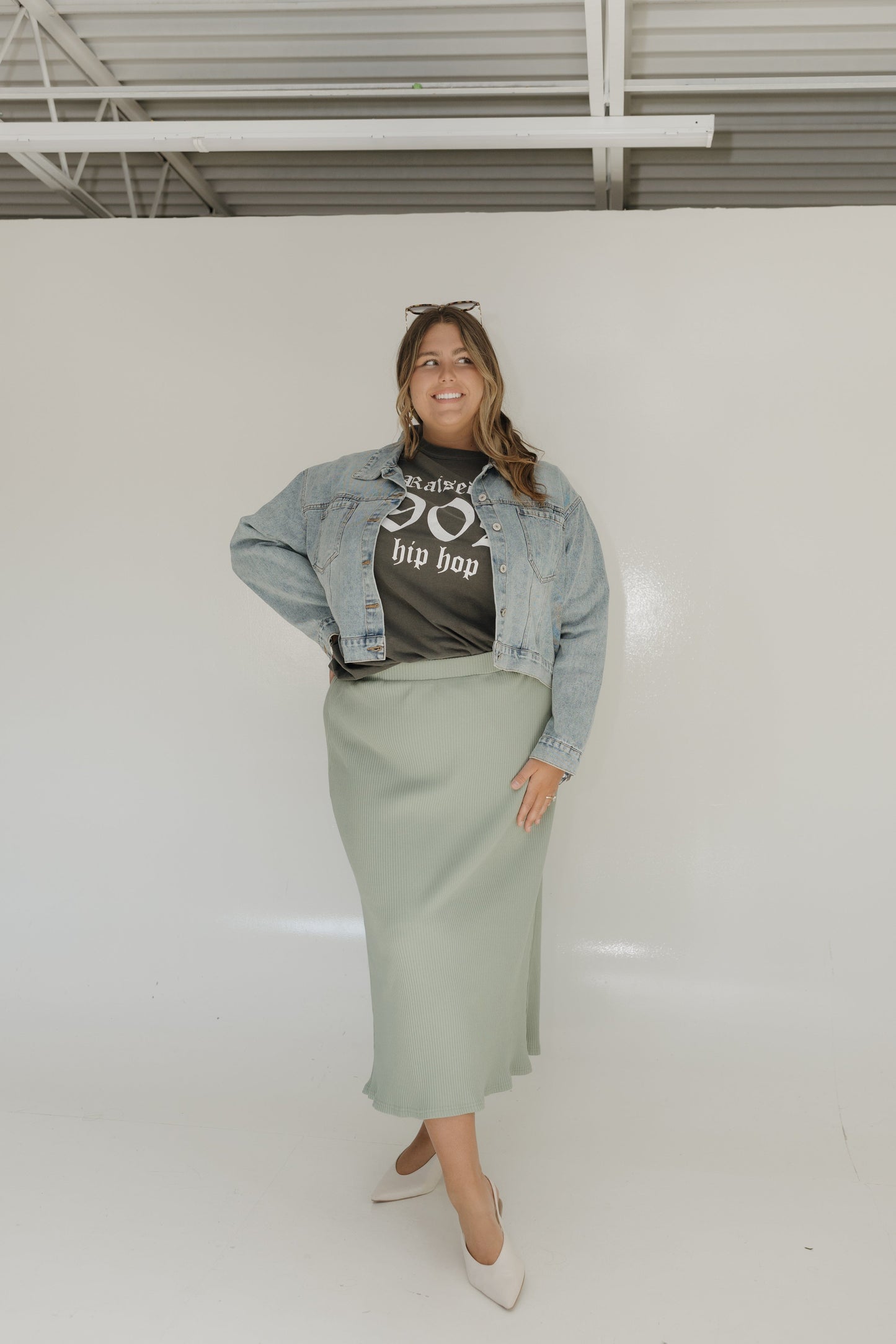 Person wearing a denim jacket, graphic t-shirt, and green skirt against a white wall.
