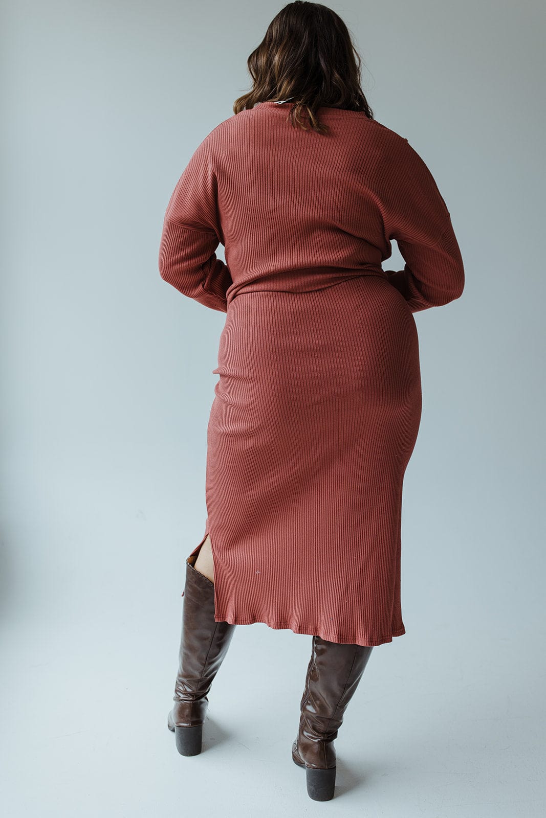 A person wearing a Mikarose ribbed knit midi-length pencil skirt in smokey rosewood, paired with tall brown boots, stands against a light grey background that evokes the subtle elegance of terracotta.