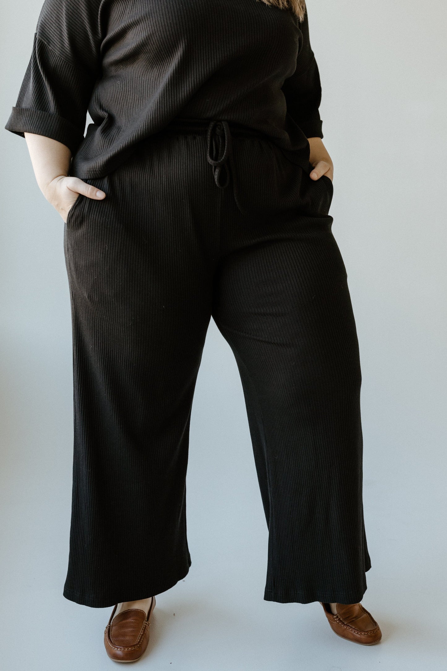 A person wearing a black ribbed outfit with high waist pants stands confidently, hands in pockets against a neutral background.