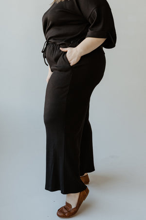 A person stands confidently in a black outfit featuring ribbed wide-leg pants, hands casually tucked in pockets. Complemented by brown shoes, they strike a stylish pose against a light gray background.