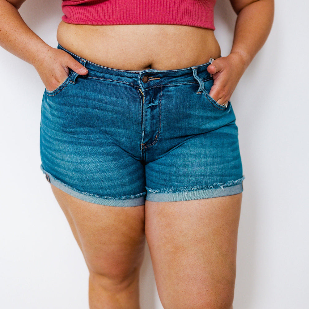 A person wearing a pink sleeveless top and Judy Blue denim shorts with a frayed hem stands with hands in pockets against a plain background.