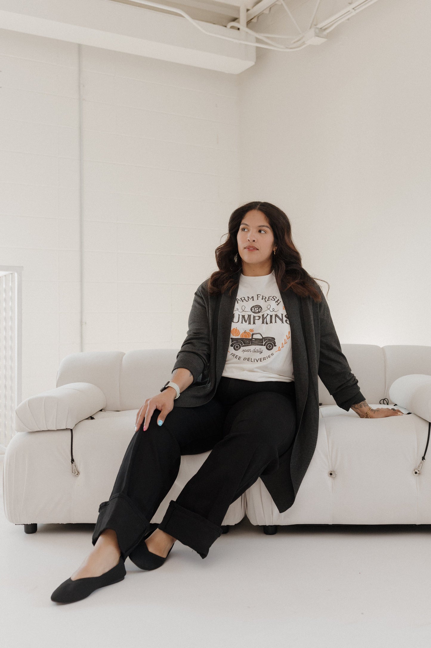 Woman sitting on a white couch wearing a graphic t-shirt and black pants in a minimalistic room.