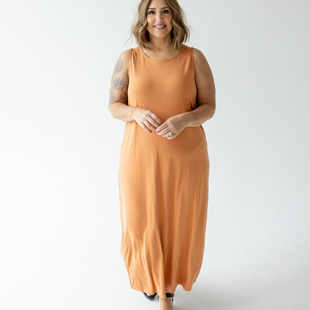 A woman with shoulder-length hair stands against a plain white background, wearing a sleeveless, floor-length, orange ROUND NECK TANK SLIP DRESS by Love Marlow and black shoes. She has a tattoo on her left arm and is smiling.