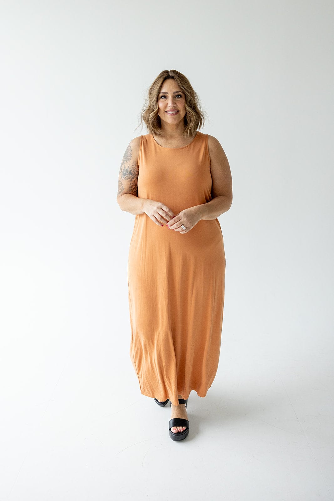 A woman with shoulder-length hair stands against a plain white background, wearing a sleeveless, floor-length, orange ROUND NECK TANK SLIP DRESS by Love Marlow and black shoes. She has a tattoo on her left arm and is smiling.