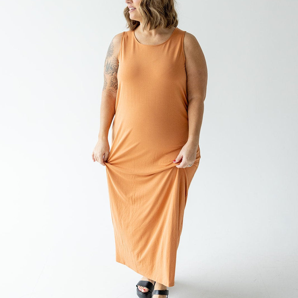 A person in an orange, ROUND NECK TANK SLIP DRESS by Love Marlow and black sandals stands against a plain white background, holding the dress with one hand and smiling.