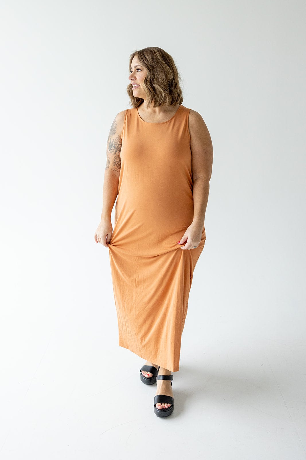 A person in an orange, ROUND NECK TANK SLIP DRESS by Love Marlow and black sandals stands against a plain white background, holding the dress with one hand and smiling.