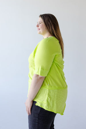 A woman with shoulder-length brown hair, wearing glasses, a ROUND NECK TUNIC WITH BUTTON DETAIL IN TENNIS GREEN from Love Marlow, and dark pants, stands facing sideways against a plain white background.