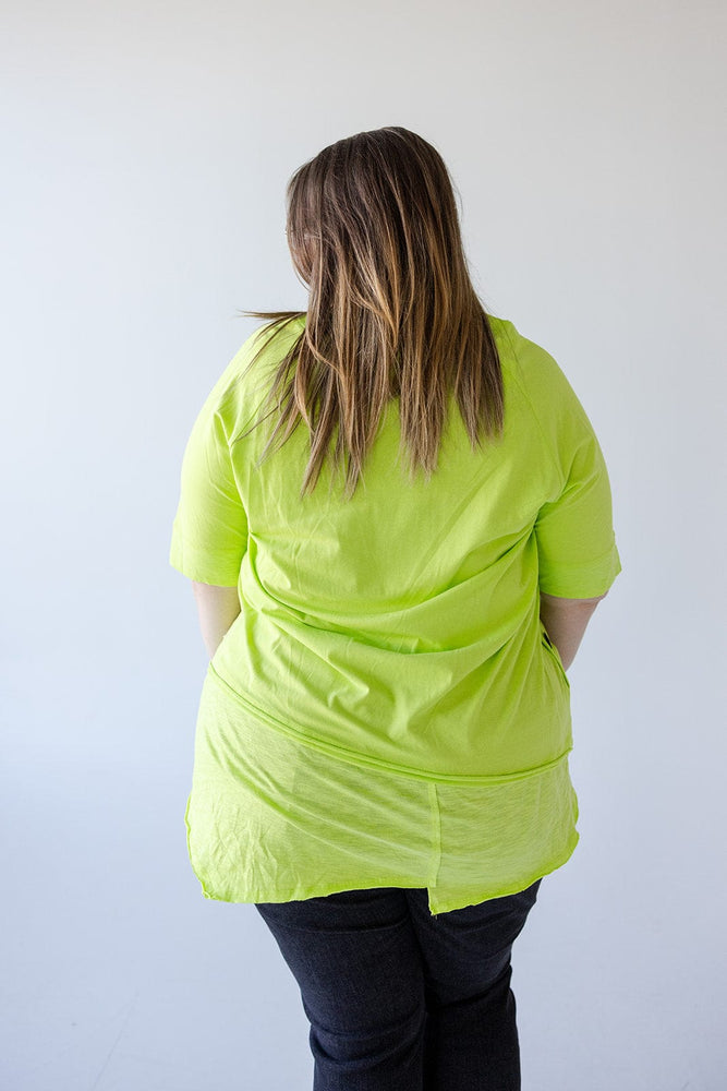 Person with shoulder-length hair, wearing a ROUND NECK TUNIC WITH BUTTON DETAIL IN TENNIS GREEN by Love Marlow and black pants made of luxurious cotton fabric, facing away from the camera in front of a plain white background.