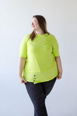 A person wearing a bright yellow-green, asymmetrical design top and dark jeans stands against a plain white background, looking to the side with a slight smile. They are donning the ROUND NECK TUNIC WITH BUTTON DETAIL IN TENNIS GREEN from Love Marlow.