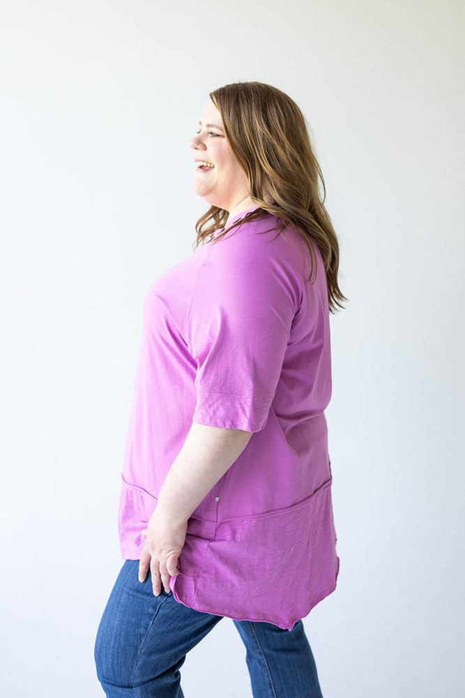 A woman with long brown hair is standing sideways, smiling, and wearing a pink Love Marlow ROUND NECK TUNIC WITH BUTTON DETAIL IN VERBENA and blue jeans against a plain white background.