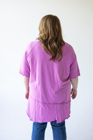 A person with shoulder-length hair wearing a pink, luxurious cotton ROUND NECK TUNIC WITH BUTTON DETAIL IN VERBENA by Love Marlow and blue jeans is standing and facing away from the camera against a plain white background.