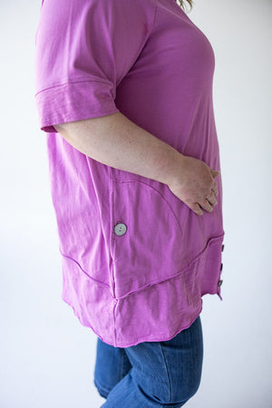 Side view of a person wearing a bright pink, Love Marlow ROUND NECK TUNIC WITH BUTTON DETAIL IN VERBENA with short sleeves and an asymmetrical hem, paired with blue jeans.