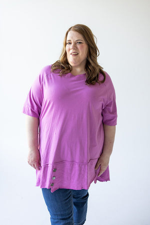 A person with long hair is wearing a pink ROUND NECK TUNIC WITH BUTTON DETAIL IN VERBENA by Love Marlow and blue jeans, standing against a plain white background.