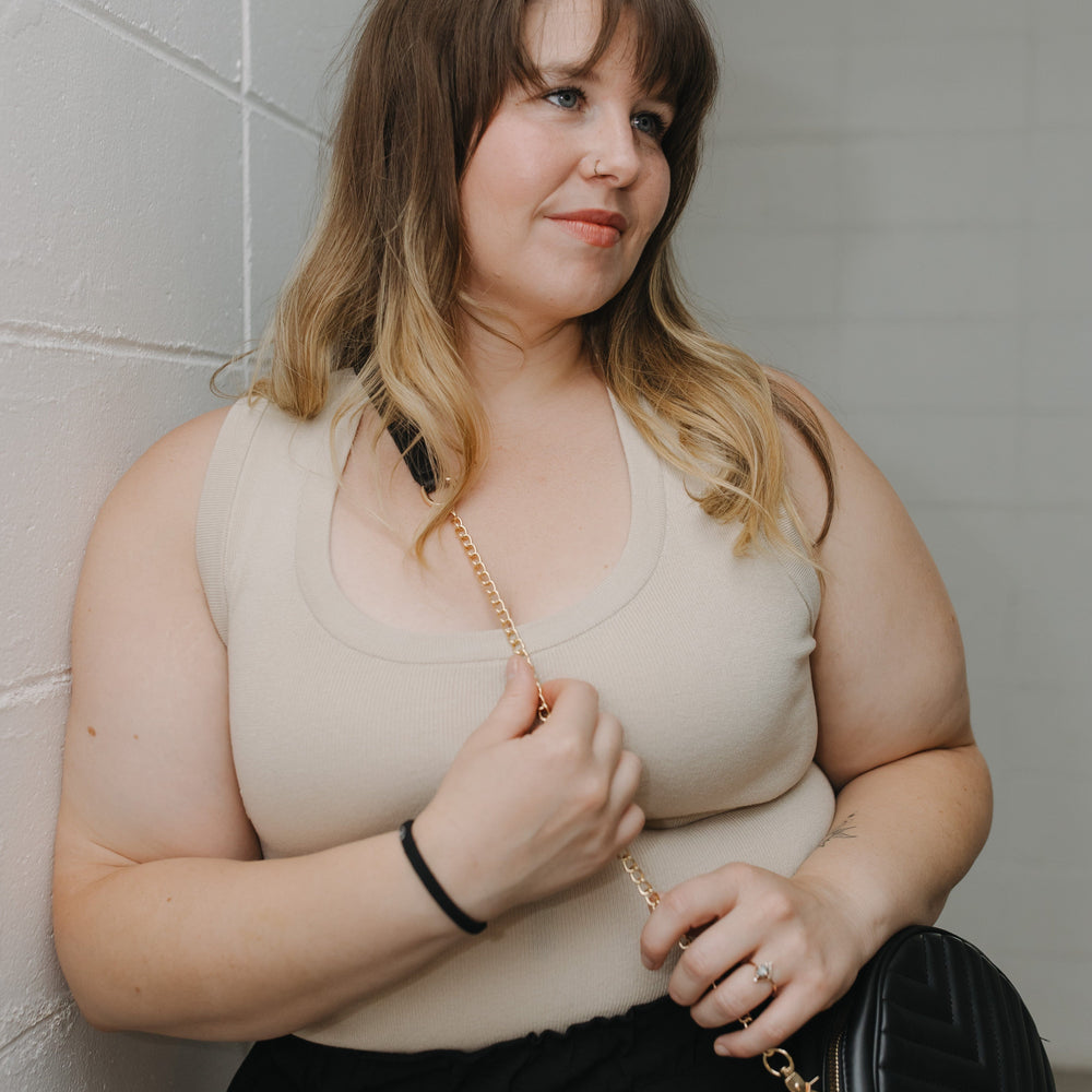 Woman holding the round quilted cross-body purse in black while she is wearing a beige top and black skirt, standing against a tiled wall.