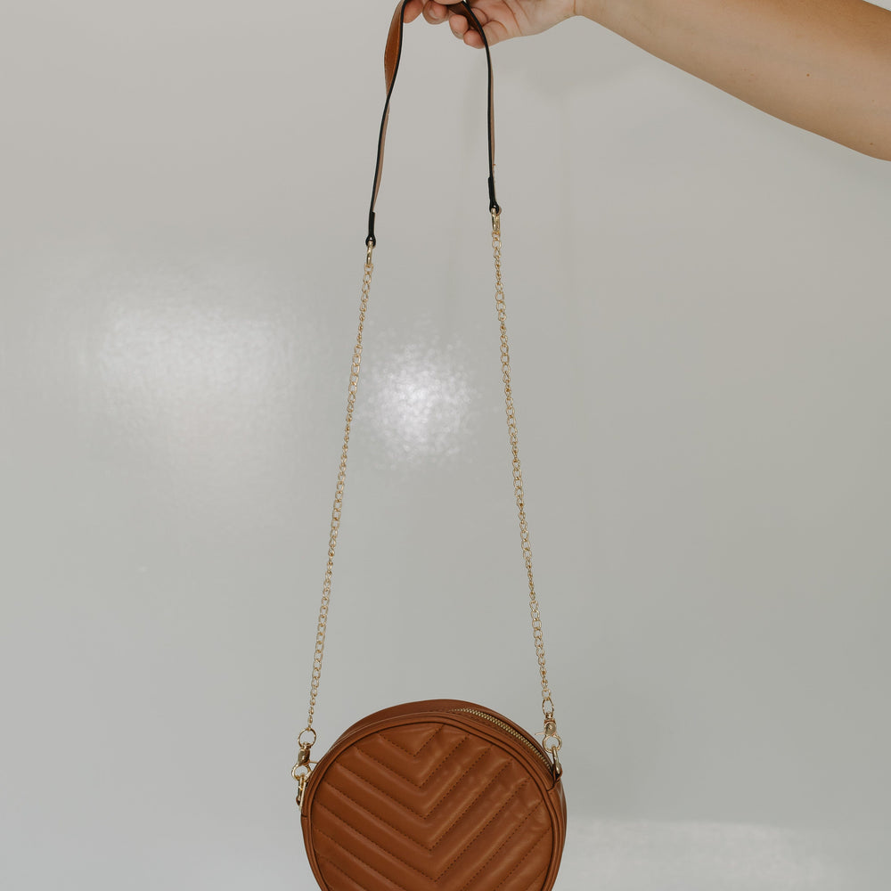 Round brown handbag with a chain strap held by a hand against a light gray background