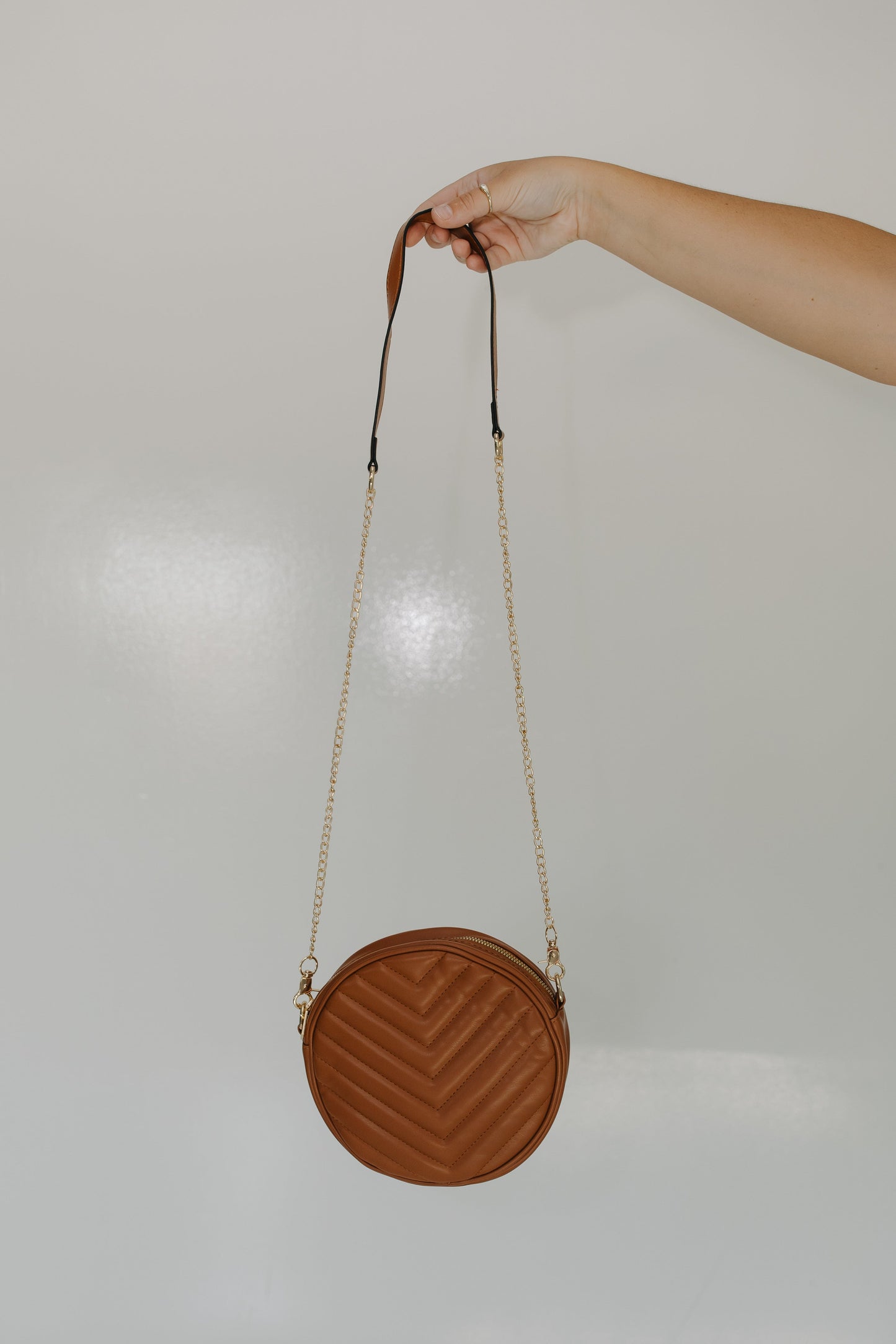 Round brown handbag with a chain strap held by a hand against a light gray background