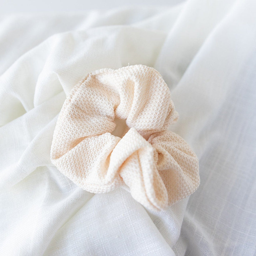 A chic, textured scrunchie in beige graces a crumpled white cloth background, creating the perfect cream hair accessory for stylish ponytails.