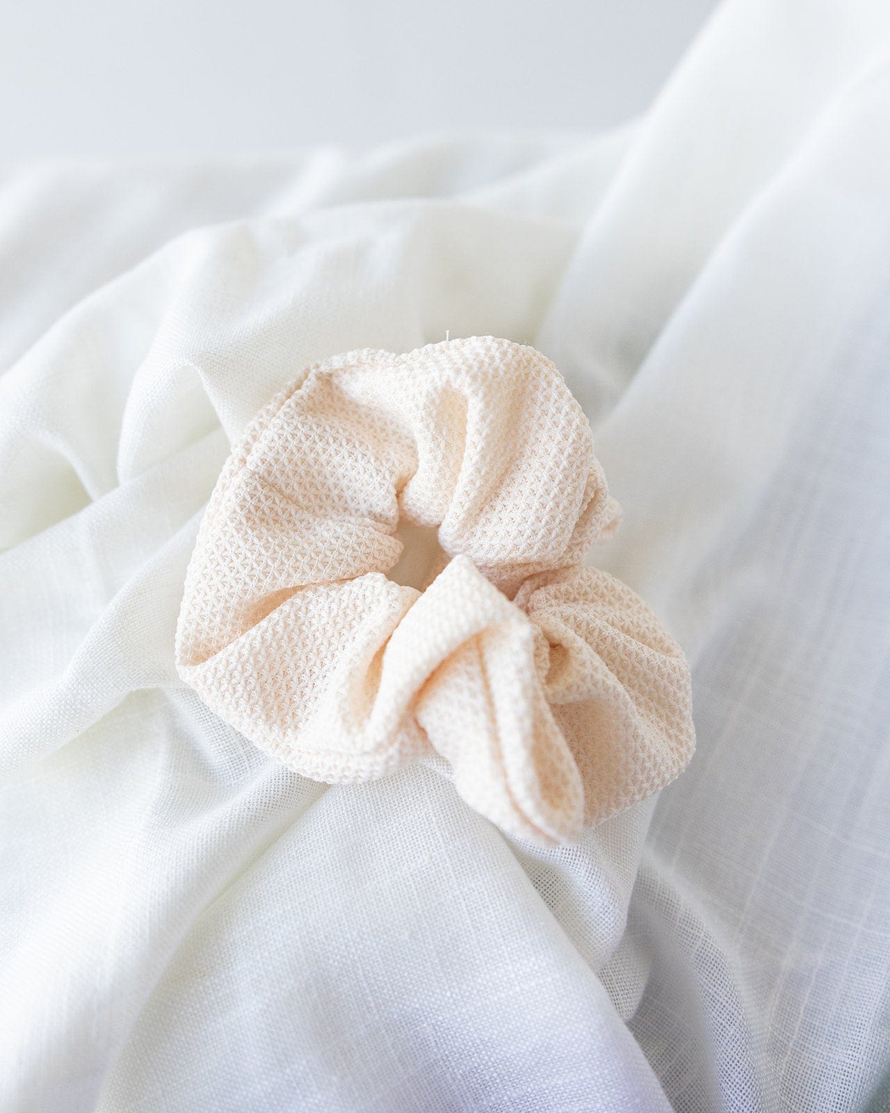 A chic, textured scrunchie in beige graces a crumpled white cloth background, creating the perfect cream hair accessory for stylish ponytails.